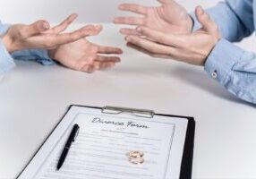 The image shows two people in light blue shirts gesturing with their hands during what appears to be a discussion about Alabama divorce, with a clipboard containing divorce paperwork, a pen, and wedding rings visible in the foreground.