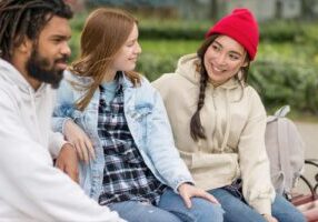 Three diverse teenagers sitting together outdoors on a bench. A young man with dreadlocks in a white hoodie is on the left, a young woman with auburn hair wearing a denim jacket and plaid shirt is in the middle, and an Asian young woman with braided hair wearing a cream-colored hoodie and red beanie is on the right.