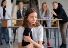 A person in the foreground appearing distressed and traumatized, while people in the background laugh, symbolizing a hostile work environment. This image relates to workplace violence, focusing on employer liability and workers' compensation for affected employees.