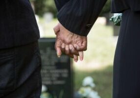 A close-up of a couple holding hands in front of a tombstone, with the stone visible in the softly blurred background. The image reflects shared grief and the bond between loved ones as they remember a life lost.